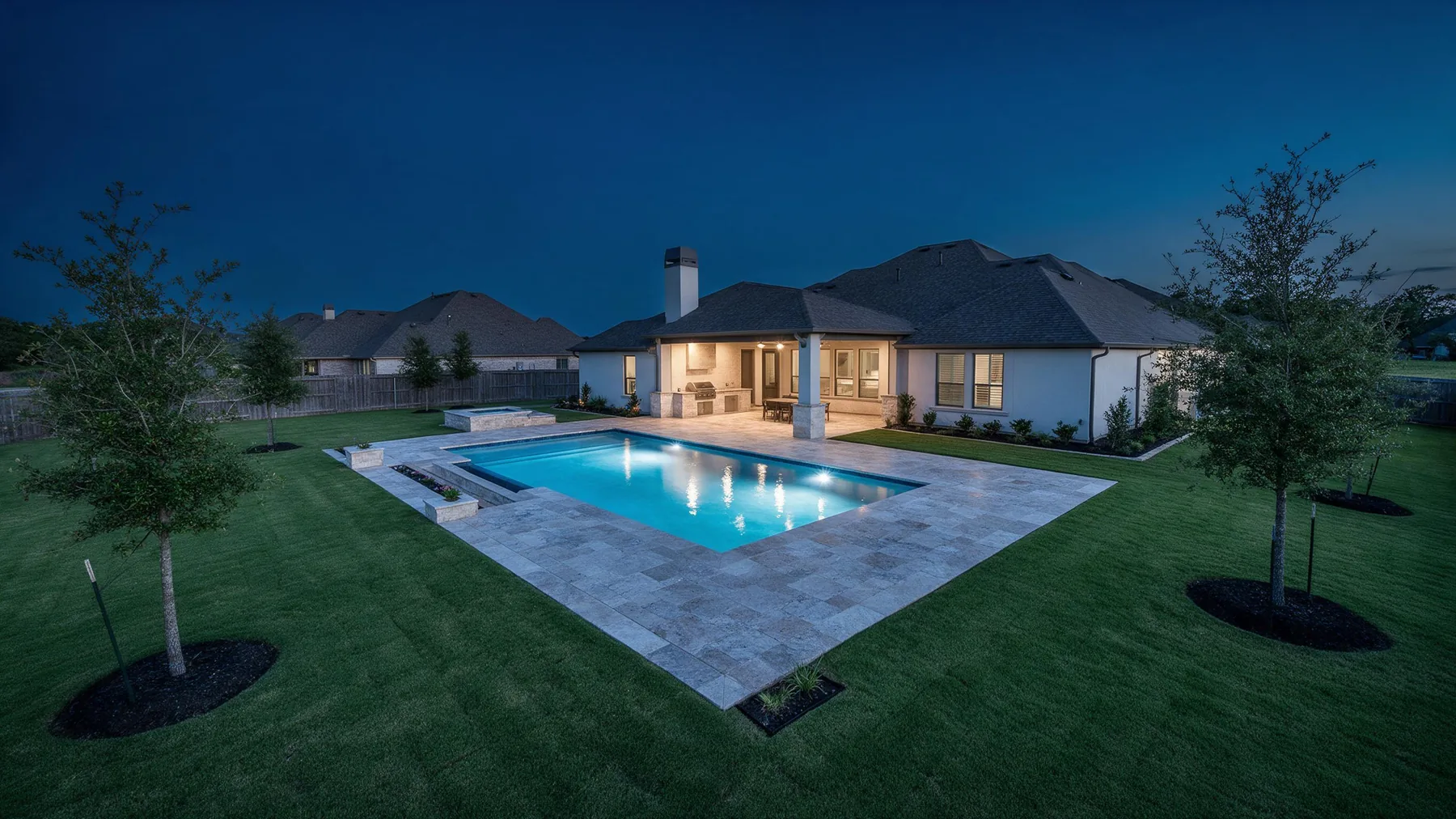 Texas home at dusk with a lit pool, travertine decking, covered outdoor kitchen and maintained lawn